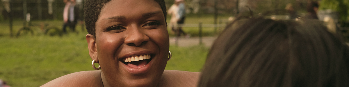 A non-binary trans woman laughing in a park.png